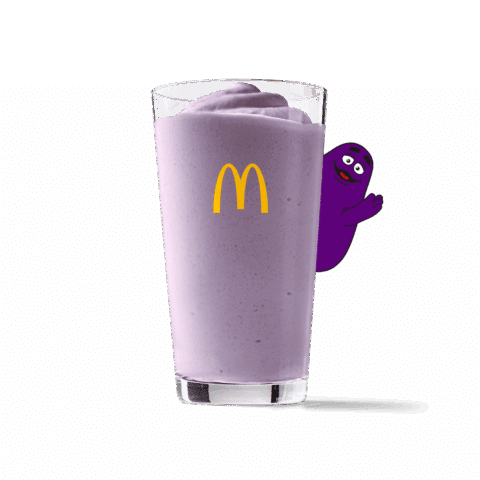 Cremiger, hellvioletter Heidelbeer-Milchshake in einem Glas mit gelbem McDonald's Logo; im Hintergrund winkt Grimace freundlich hinter dem Glas hervor.