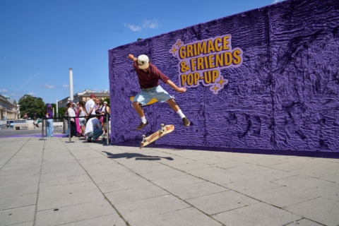 Skater springt über eine Skateboardrampe vor einer lilafarbenen Wand mit der Aufschrift "Grimace & Friends Pop-Up"