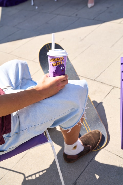 Eine Hand hält einen Trinkbecher mit dem Schriftzug 'Grimace Shake', während die Person auf einem Skateboard sitzt.