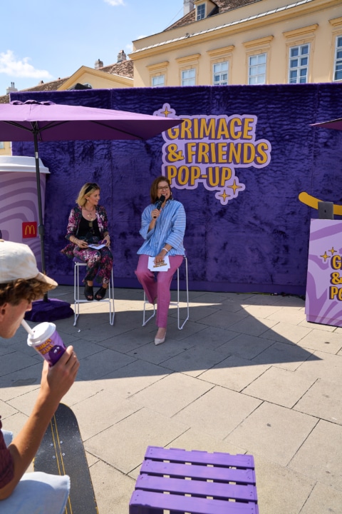 Zwei Frauen auf einer Bühne beim Grimace & Friends Pop-Up Event, umgeben von lila Farbtönen und einer McDonald's-Einrichtung.
