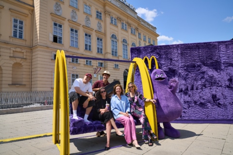 Gruppe von fünf Menschen, die auf einer schwingenden Bank mit dem McDonald's-Maskottchen Grimace posieren, vor einem historischen Gebäude an einem sonnigen Tag.