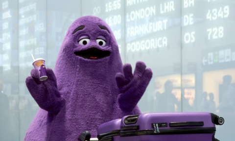 Lila, plüschige Figur mit einem Getränk in der Hand, steht vor einem Flughafen-Anzeigeboard.