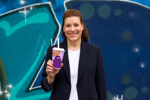 Frau in schwarzem Blazer hält einen Getränkebecher mit Grimace Shake im Vordergrund einer grafischen Wandmalerei.