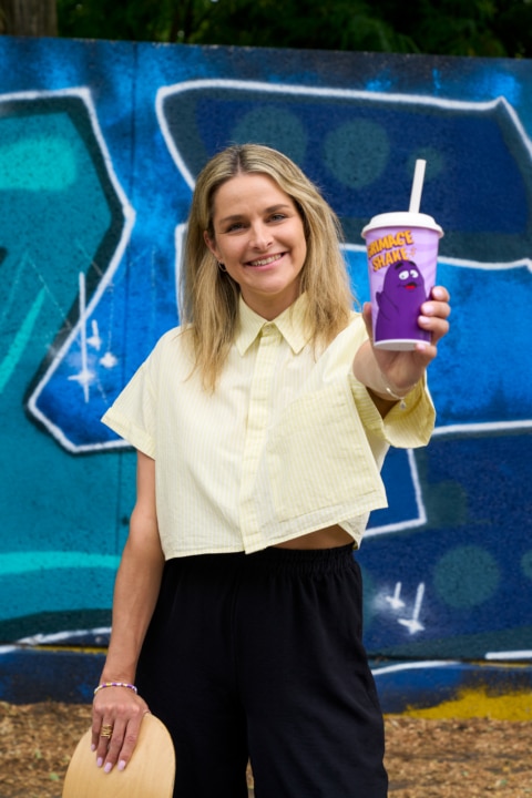 Junge Frau mit blonden Haaren, die ein Getränk in einer lila Becher präsentiert, vor einer bunten Graffiti-Wand.