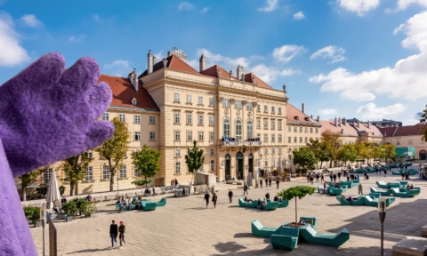 Blick auf den Platz vor einem historischen Gebäude, umgeben von Menschen und modernen Sitzgelegenheiten