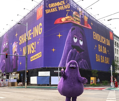 Maskottchen Grimace von McDonald's steht vor einer großen Werbetafel, die das Grimace Shake Menü bewirbt.