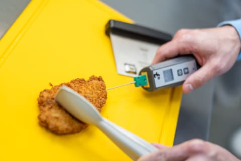 Ein Koch misst die Temperatur eines panierten Hähnchens mit einem digitalen Thermometer auf einem gelben Schneidebrett.
