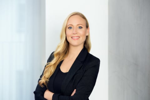 Professionelle Frau mit langen blonden Haaren in einem schwarzen Blazer, die lächelt und mit verschränkten Armen vor einer hellen Wand steht.
